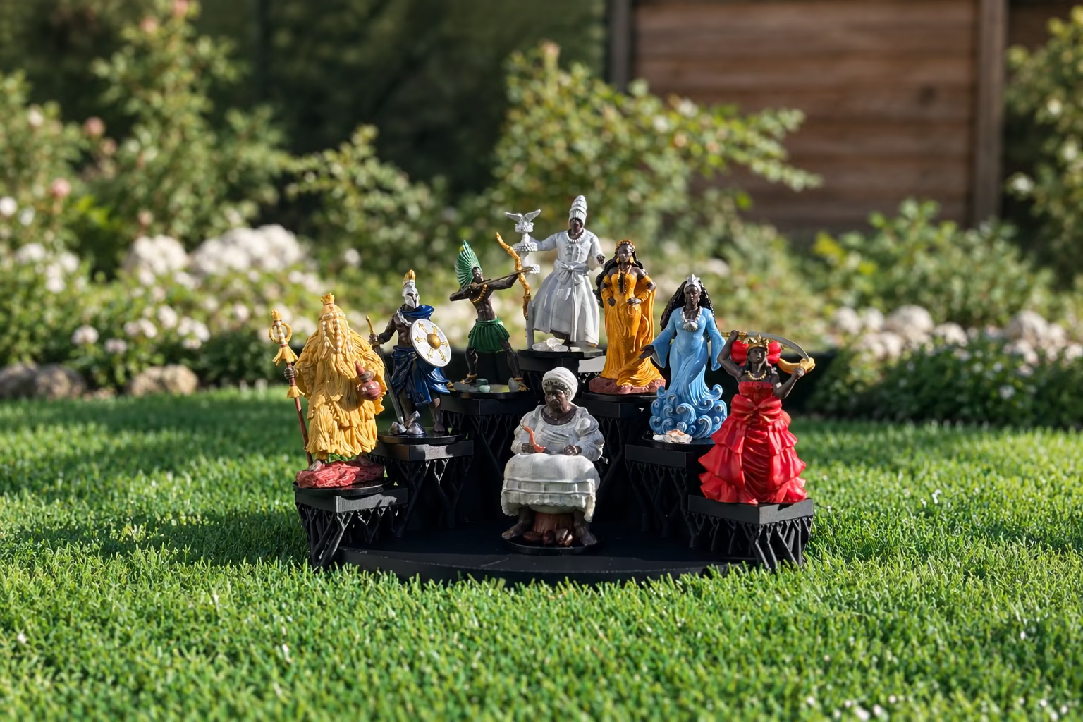 A set of nine colorful African royal and historical figurines displayed on a black multi-tiered base in an outdoor garden setting.